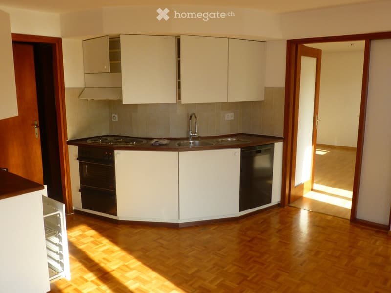 Modern kitchen with white cabinets, wooden flooring, and built-in appliances including an oven, dishwasher, and refrigerator. The space is well-lit with natural light streaming in from an adjacent room.