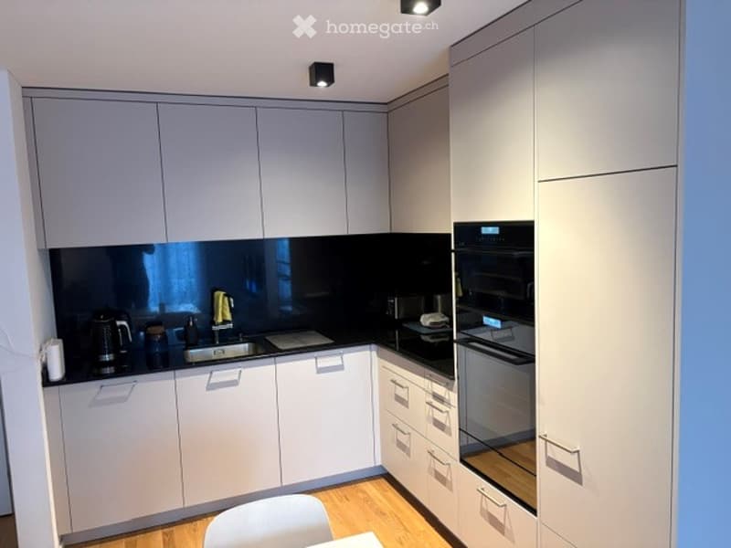Modern kitchen with white cabinets, black countertops, and stainless steel appliances including an oven and coffee maker. A white chair is visible in the foreground.