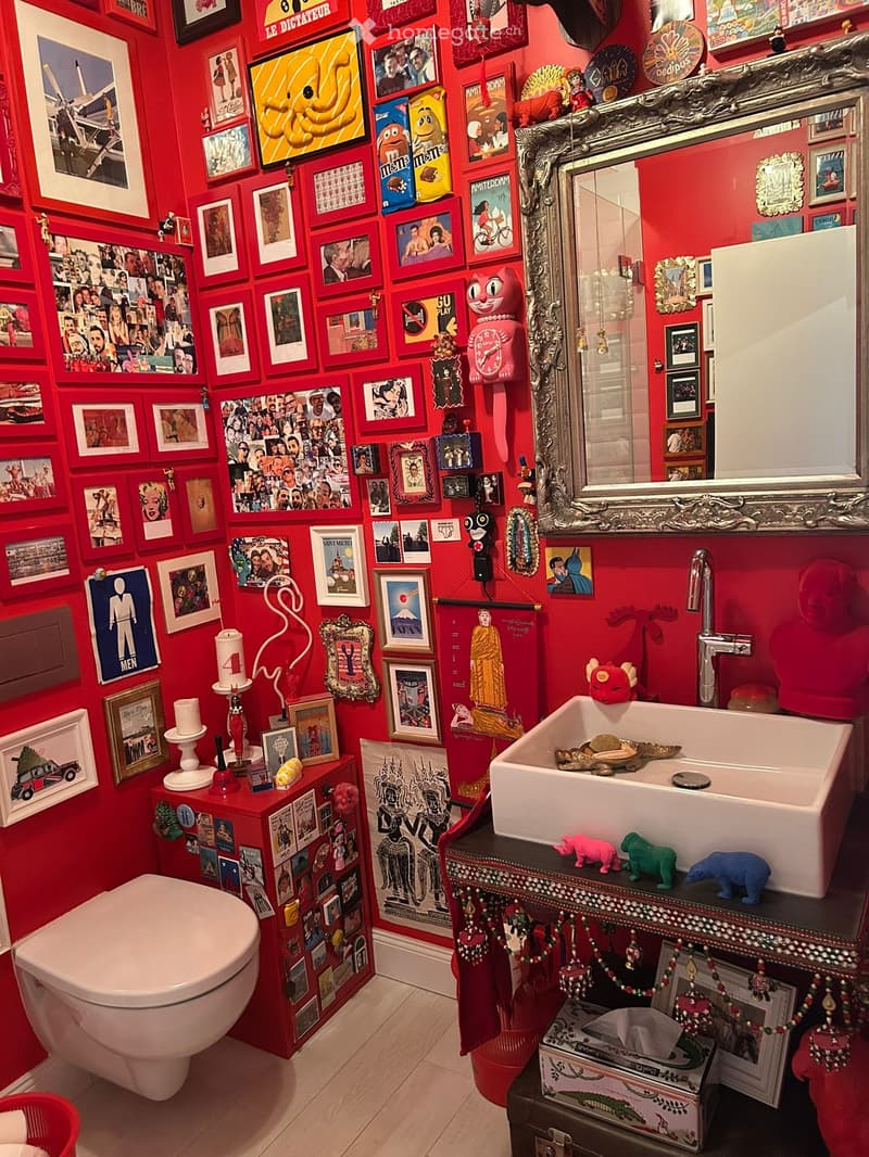 A vibrant red bathroom with a toilet, sink, and ornate mirror. The walls are covered with numerous framed pictures and posters, creating a colorful and eclectic atmosphere. A small cabinet holds various decorative items, including a clock, candle, and sma