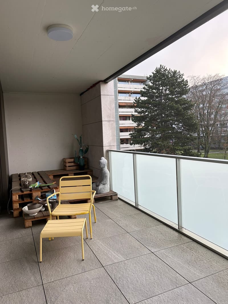 Balcony with modern glass railing, tiled floor, yellow chair and stool, small table with decorative items, statue, and potted plants.