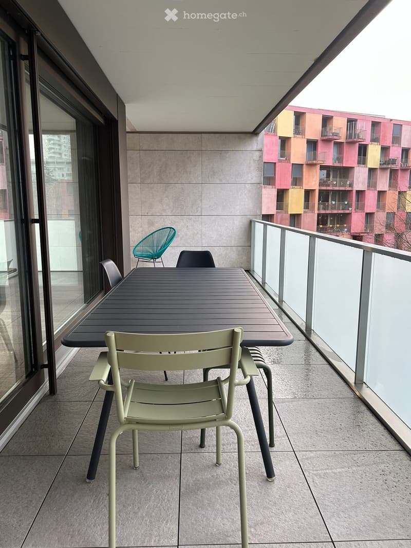 Balcony with a dark wooden table and four chairs, including a green and a teal chair, overlooking a colorful residential building.