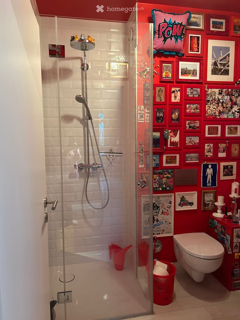 Modern bathroom with glass shower enclosure, white tiles, red walls, decorative pictures, white toilet, red storage box, red bucket, and shower head.