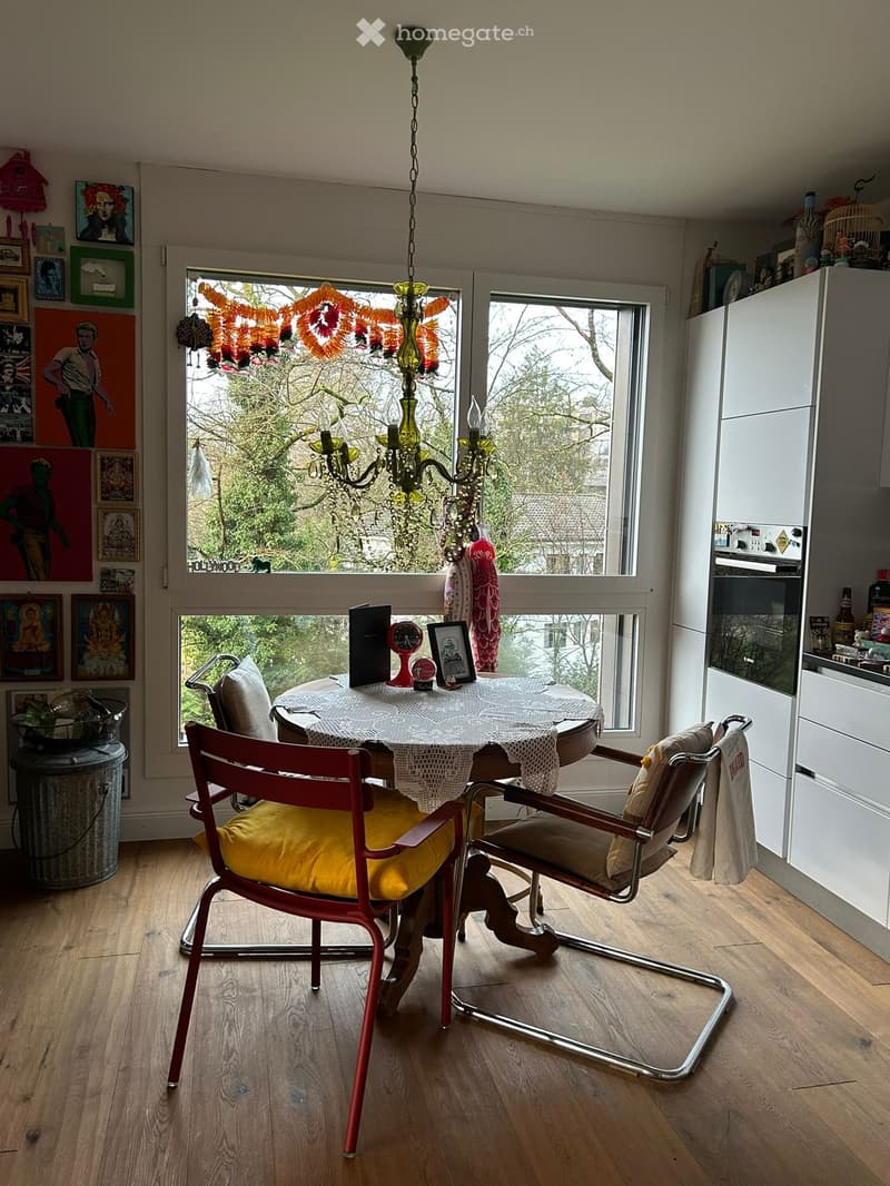 Dining room with a round table, chairs, a chandelier, and a window with a view of trees outside.