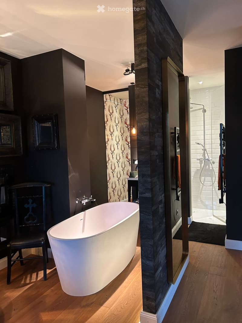 Modern bathroom with a freestanding bathtub, dark walls, wooden flooring, a glass-enclosed shower, and a small table with a mirror.