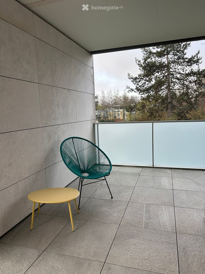 Modern balcony with a green wire chair and a yellow round table, overlooking a tree-lined area.