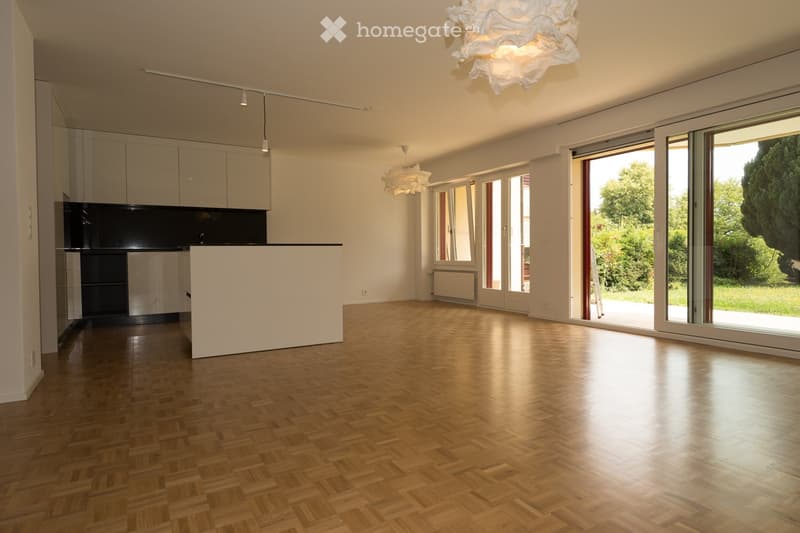 Open-plan unfurnished room with light wooden flooring, white walls, modern kitchen area with cabinets and a countertop, large windows and sliding glass doors leading to an outdoor area, and contemporary pendant lights.
