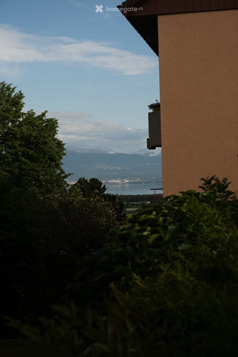 Balcony with a scenic view of mountains, a lake, and a town in the distance, surrounded by greenery.