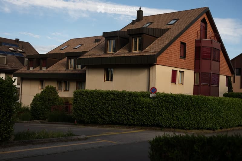 Two-story house with a brown tiled roof, multiple windows, and a red balcony section. The house is surrounded by greenery and a hedge, with a parking area in front.