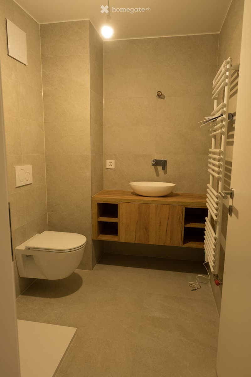 Modern bathroom with beige tiles, a toilet, a wooden cabinet with a sink, and a white radiator on the wall.