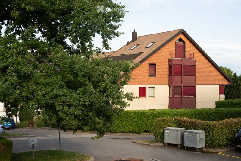 Two-story house with a red-tiled roof, beige walls, and red shutters. Features include a balcony on the upper floor and a well-maintained hedge in the front yard.