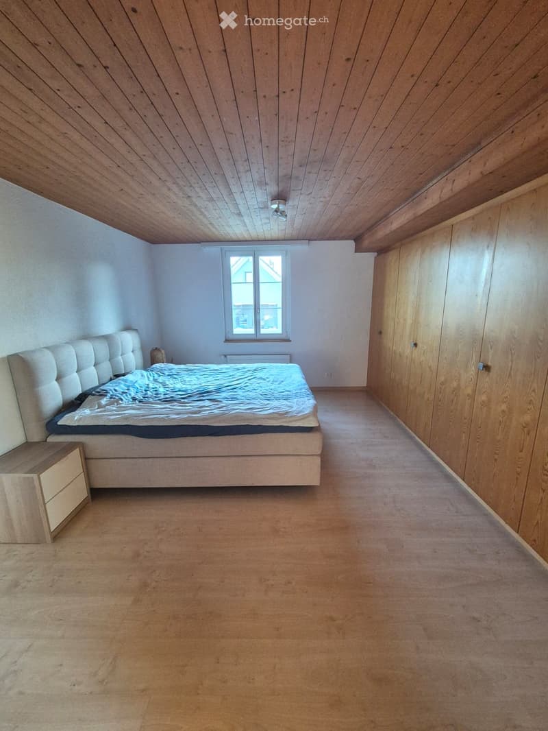Bedroom with wooden floor, slanted wooden ceiling, large window, bed with white headboard, nightstand, and built-in wardrobe.