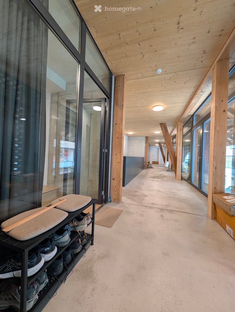 Modern hallway with wooden ceiling, concrete floor, large glass windows, and a shoe rack with shoes and a cushion on the left side.