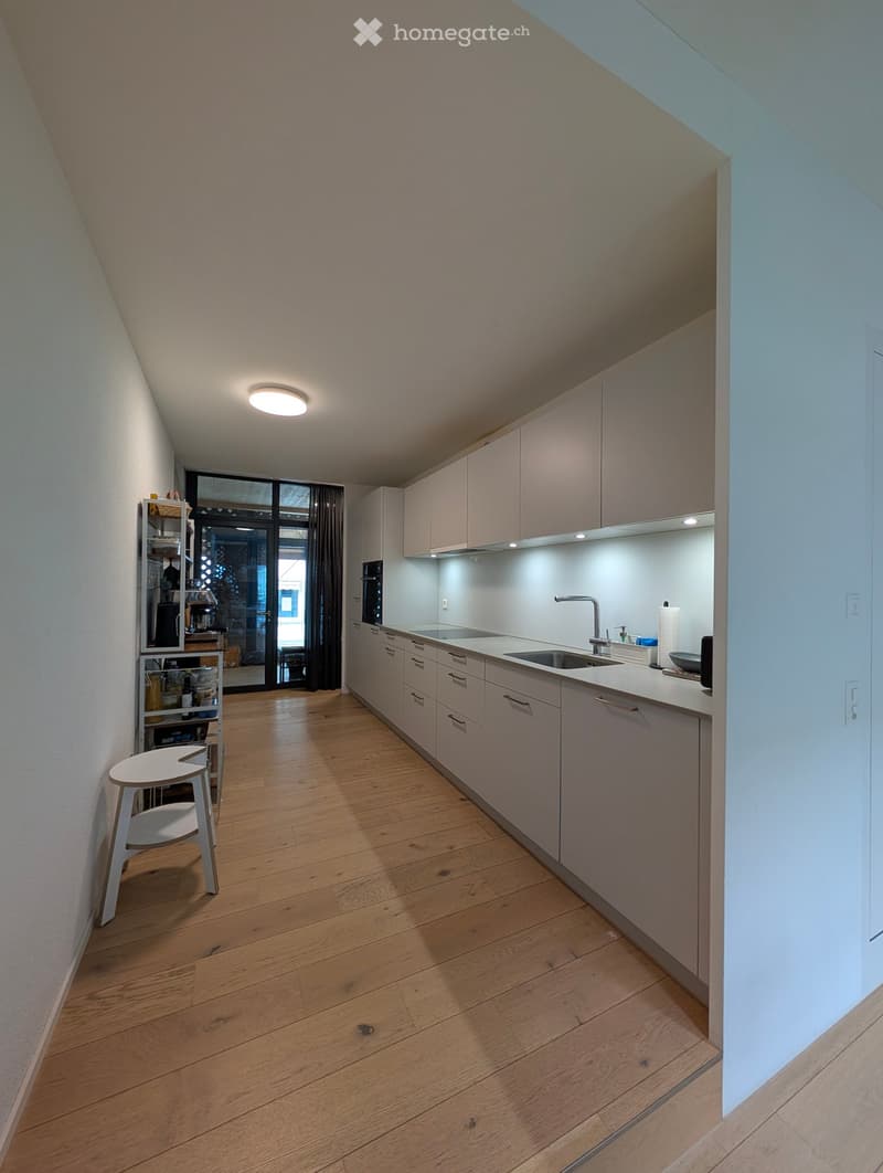 Modern kitchen with light-colored cabinets, a sink, and a small white stool near a shelf. The floor is made of light wooden planks, and the space is well-lit with recessed lighting.