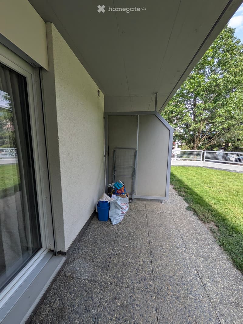 Balcony with tiled floor, white walls, and a view of a grassy area with trees.