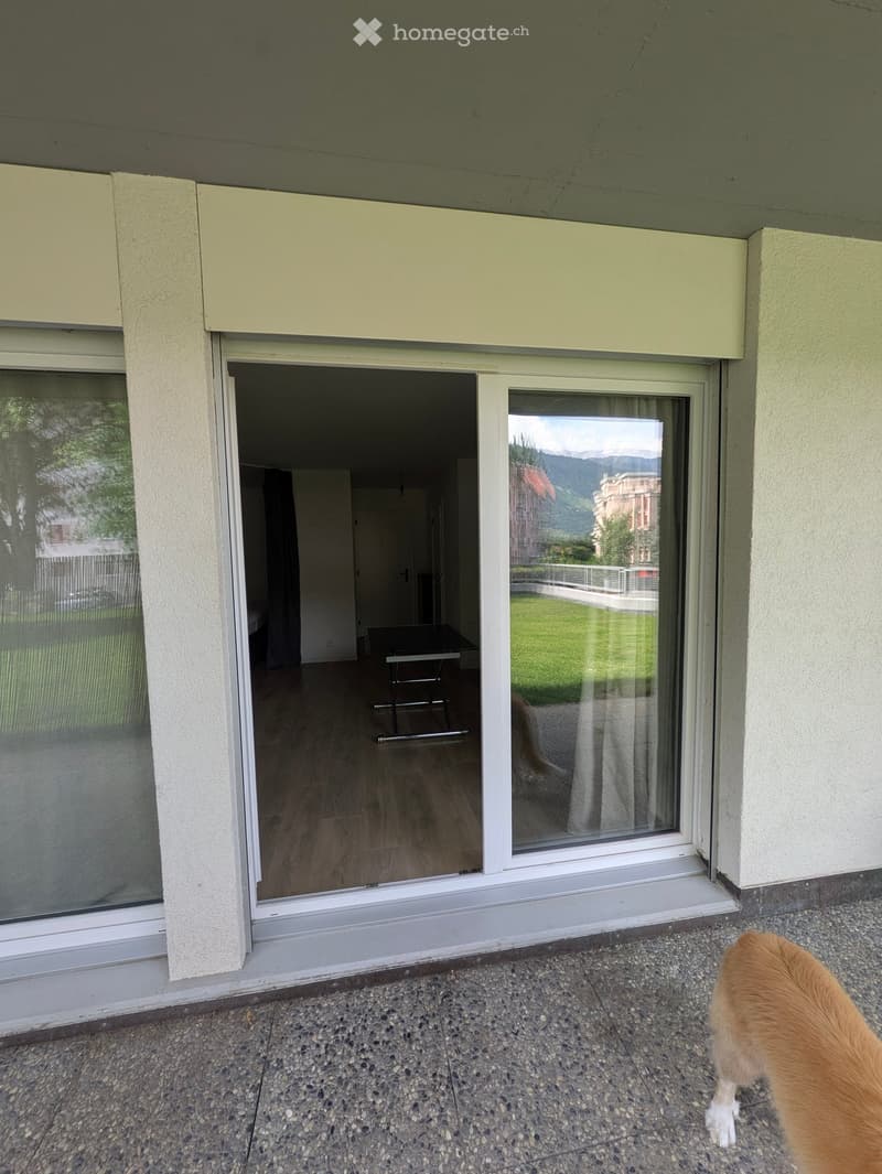 Balcony with sliding glass doors leading to a room with wooden flooring and a small table. The view shows a green lawn and distant mountains.