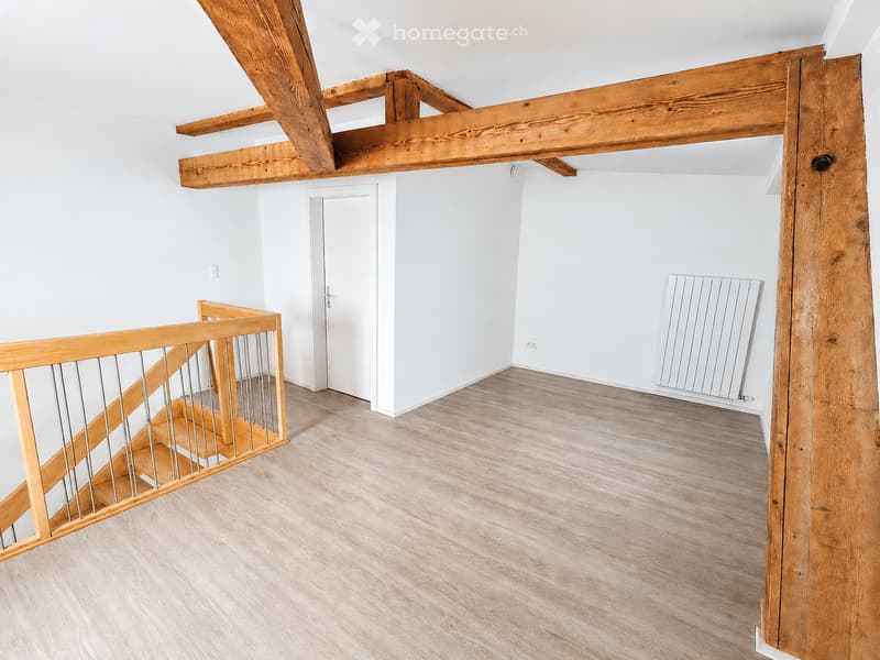 Empty room with exposed wooden beams, light wooden flooring, a staircase with a wooden railing, and a white door leading to another area.
