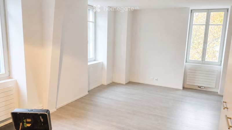 Empty room with light-colored walls, wooden flooring, and large windows. No furniture or equipment visible.
