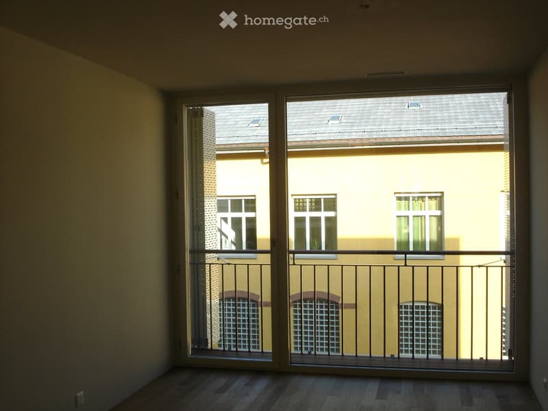 Empty room with wooden flooring, large windows, and a balcony overlooking a neighboring building.