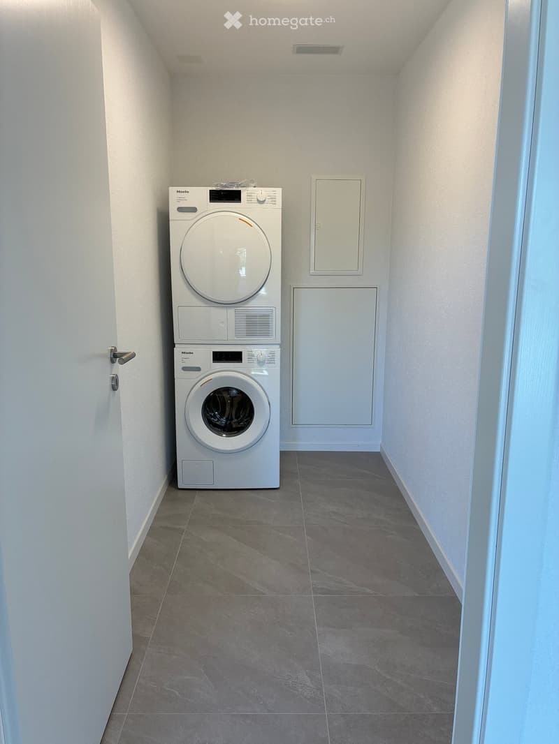 A narrow hallway with a washing machine and dryer unit against the wall, grey tiled floor, white walls, and a door on the left side.