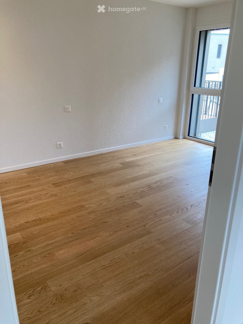 Empty room with light wooden flooring and white walls, featuring a sliding glass door leading to a balcony.
