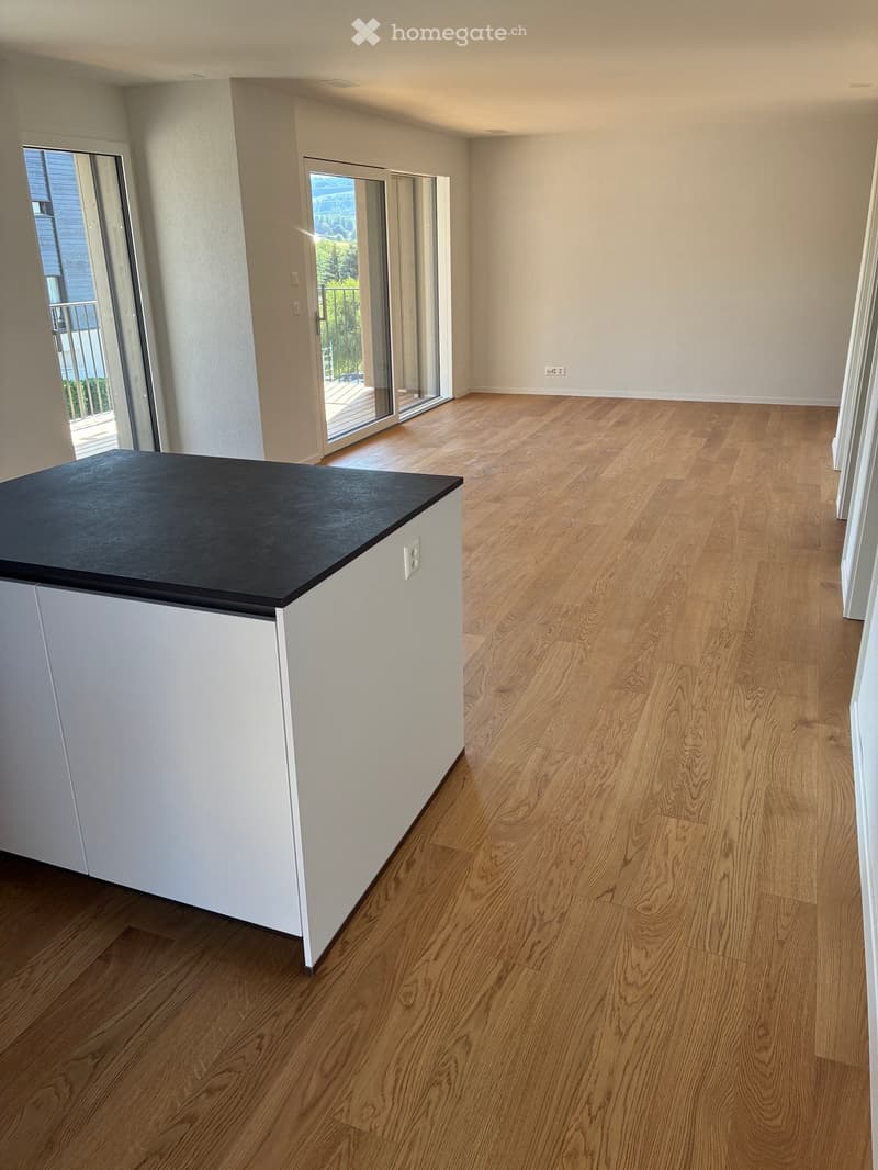Empty room with light wooden flooring, white walls, and a small white cabinet with a black countertop. There is a large sliding glass door leading to a balcony with a view of greenery.