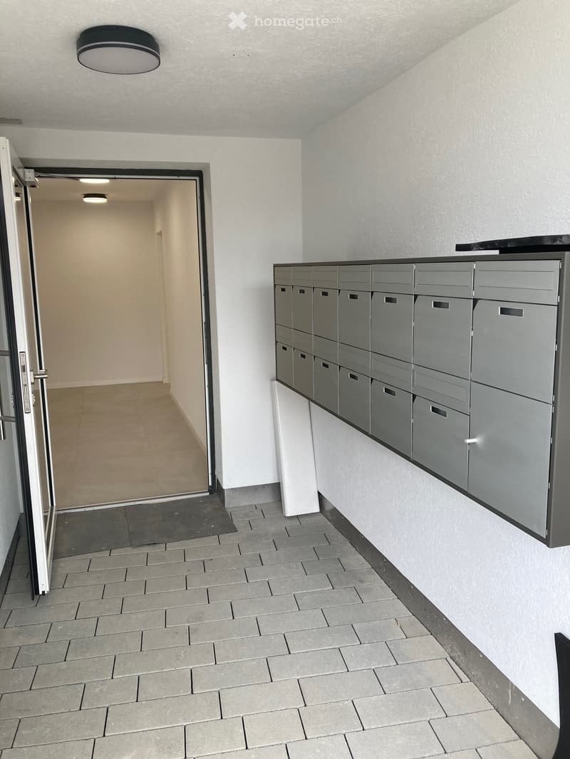 Empty hallway with gray tiled floor, white walls, and a row of mailboxes on the right side. An open door leads to another area with beige flooring.