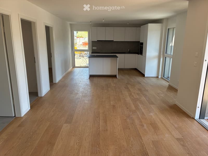 Empty room with light wooden flooring, white walls, and a small kitchen area visible in the background.