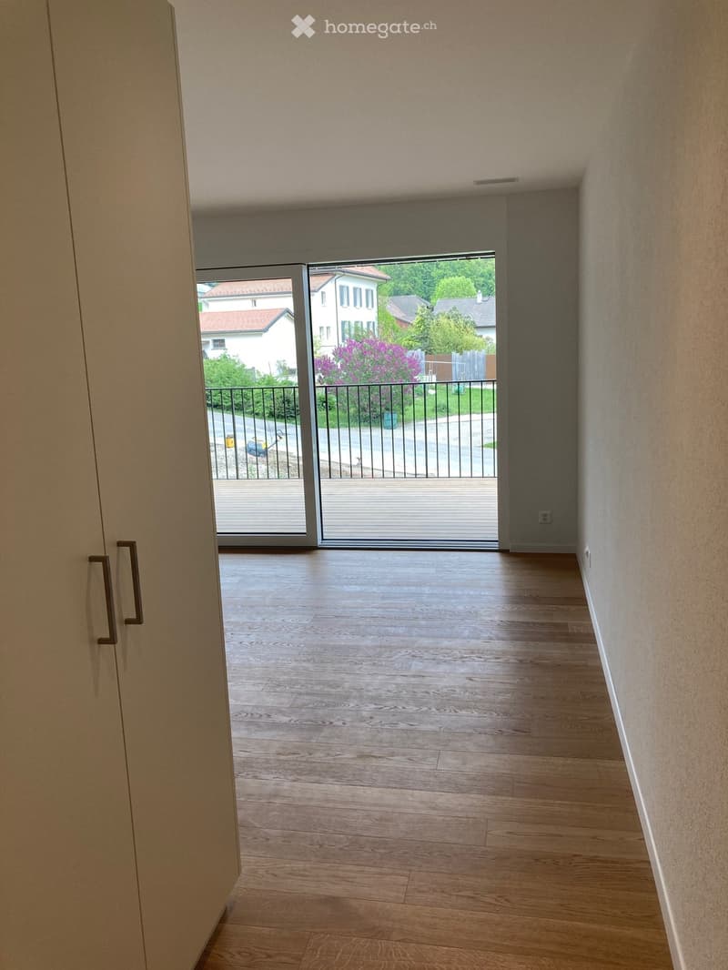 Empty hallway with wooden flooring, white walls, and a sliding glass door leading to a balcony with a metal railing and outdoor view.