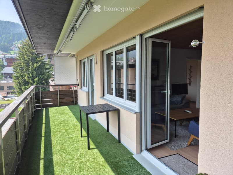 Balcony with artificial grass, small table, and railing, overlooking a scenic mountain view. Open sliding doors lead to a living room with a sofa, coffee table, and TV.