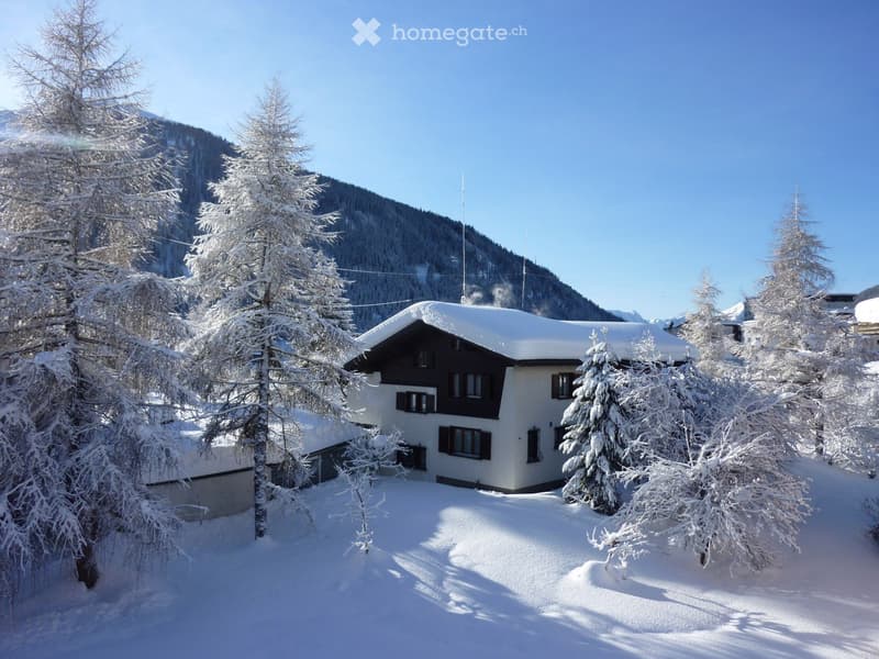 A two-story house with a snow-covered roof and white walls, surrounded by snow-laden trees and a mountainous backdrop.