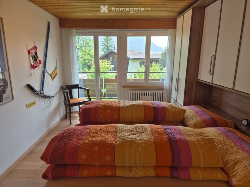 Cozy bedroom with two single beds, colorful striped bedspreads, a wooden chair, a radiator, and a built-in cabinet. Large windows offer a scenic view of the surrounding landscape.