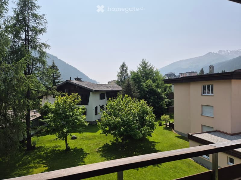 View from a balcony showing a well-maintained lawn, trees, and neighboring houses with mountain backdrop.