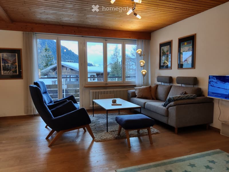 Modern living room with wooden floors, large windows, a grey sofa, coffee table, two armchairs, ottoman, and a television. The room features a cozy atmosphere with warm lighting and scenic mountain views.