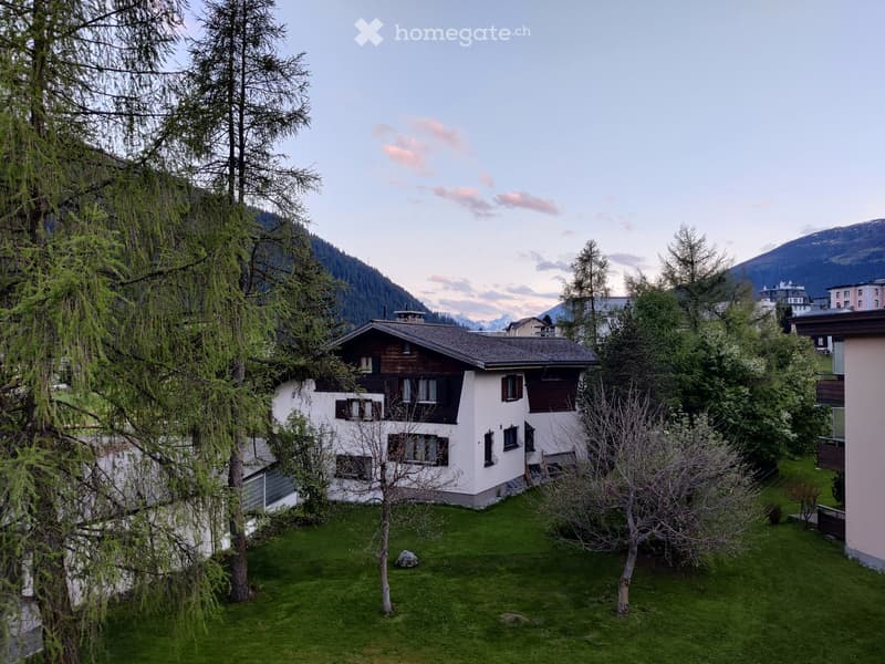 A two-story house with white walls and dark wooden shutters, surrounded by trees and a grassy lawn, with mountains in the background.