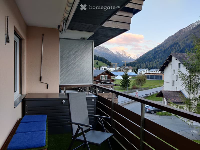Balcony with a view of mountains, featuring a folding chair and blue stools, with a railing and a small storage unit.