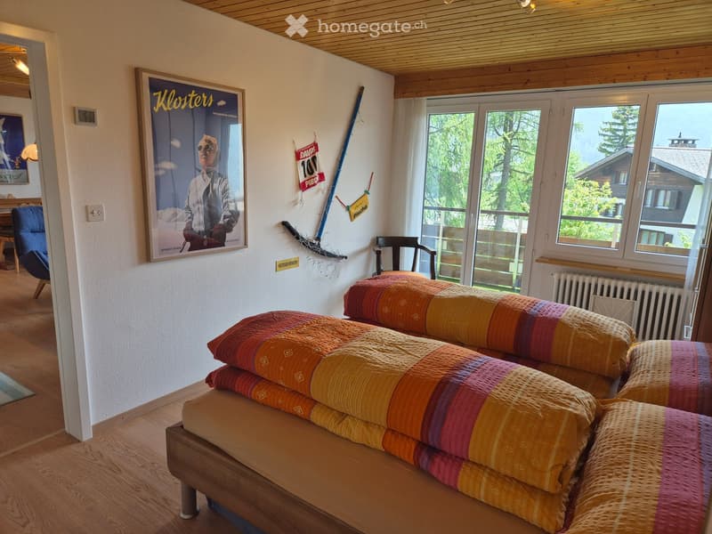 Cozy bedroom with wooden floors, a large bed with colorful bedding, a chair, a sofa, and a poster on the wall. Large windows offer a scenic view of the outdoors.