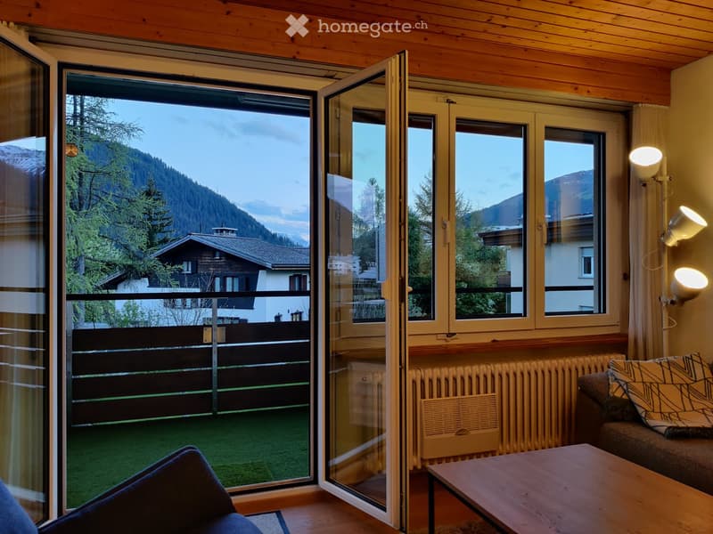Living room with wooden ceiling, large windows, open sliding doors leading to a balcony, sofa, coffee table, and a view of mountains and trees.