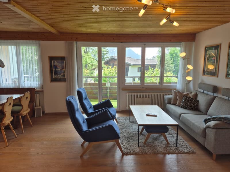 Bright living room with wooden floors, large windows, a grey sofa, coffee table, and blue armchairs.