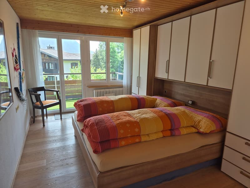 Bright bedroom with wooden floors, large windows, and a bed with colorful bedding. Includes a chair and a wardrobe.