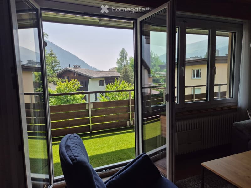 A modern living room with large windows offering a scenic mountain view, featuring a sofa and a wooden table.