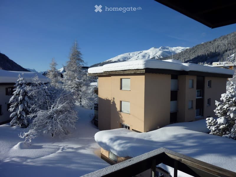 A snow-covered balcony overlooking a snow-covered building and surrounding snowy landscape with trees and mountains.