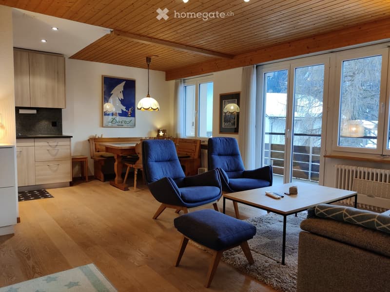 A cozy living room with wooden floors, a slanted wooden ceiling, and large windows. Features include a sofa, coffee table, chairs, a small dining area, and a kitchen area with cabinets and a refrigerator.