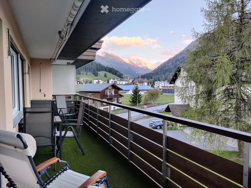 Balcony with wooden railing, green artificial turf, several chairs and a small table, overlooking a scenic mountain landscape.