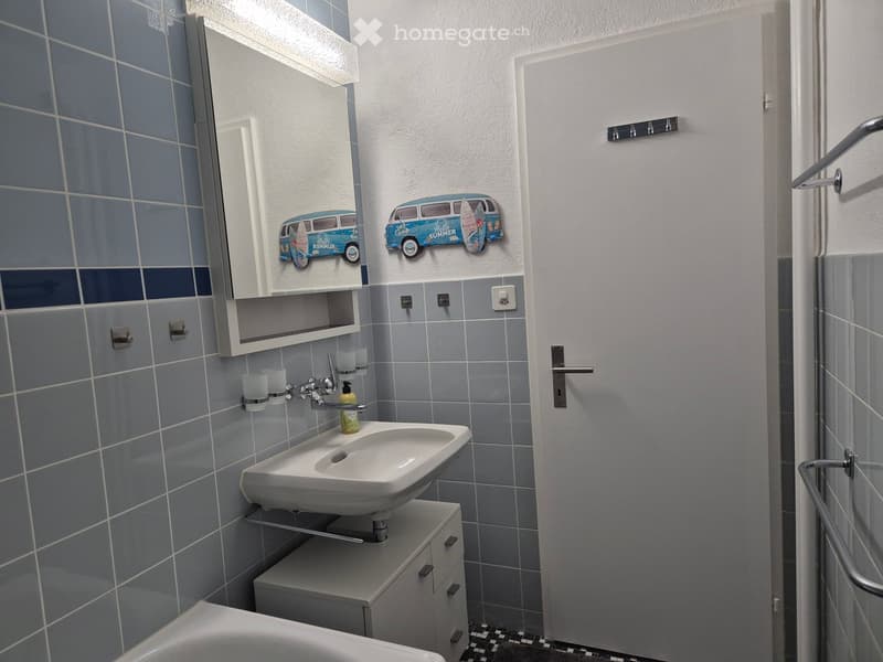 Modern bathroom with a white sink, mirror, cabinet, and bathtub. The walls are tiled in blue and grey, and there are decorative van stickers on the wall.