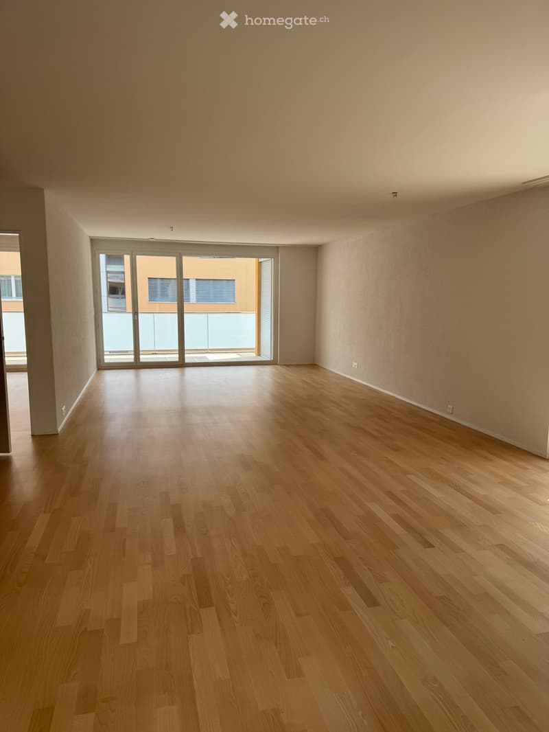 Empty room with light wooden flooring and large sliding glass doors leading to a balcony or outdoor area. The walls are painted a light beige color, and the room has a clean, minimalist design with no furniture or decorations.