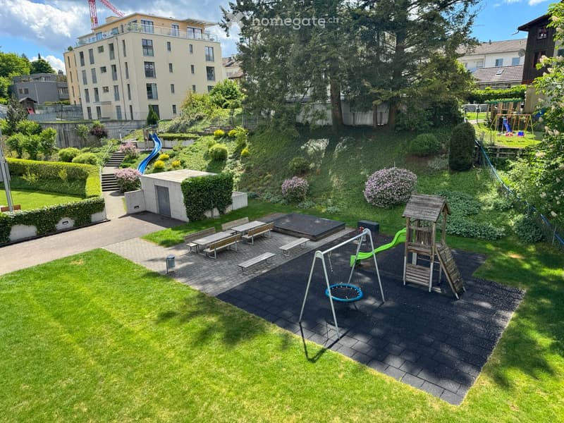 Well-maintained garden area with a playground, benches, a slide, swings, and a small wooden playhouse. The area is surrounded by greenery and includes a paved walkway and a small concrete structure.