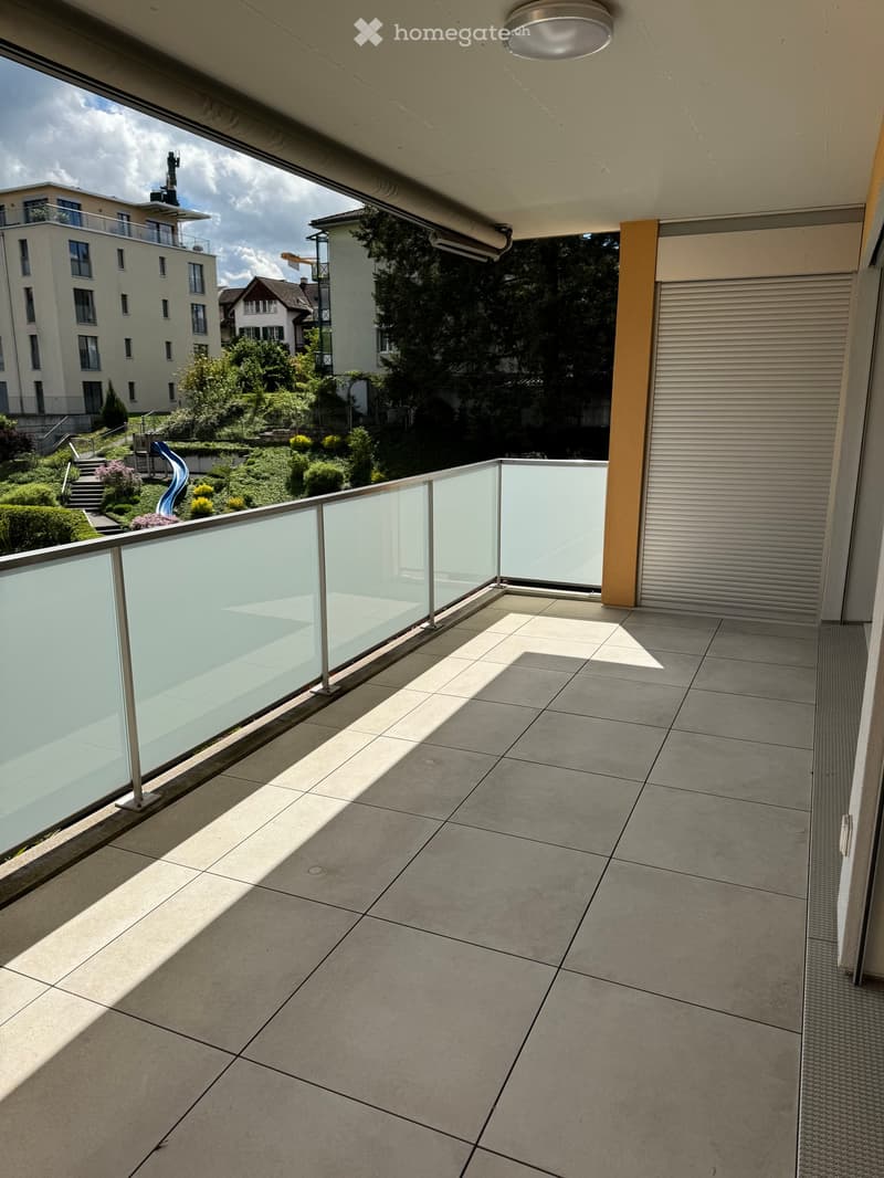 Modern balcony with grey tiled floor, glass railing, and view of a landscaped garden and neighboring buildings.