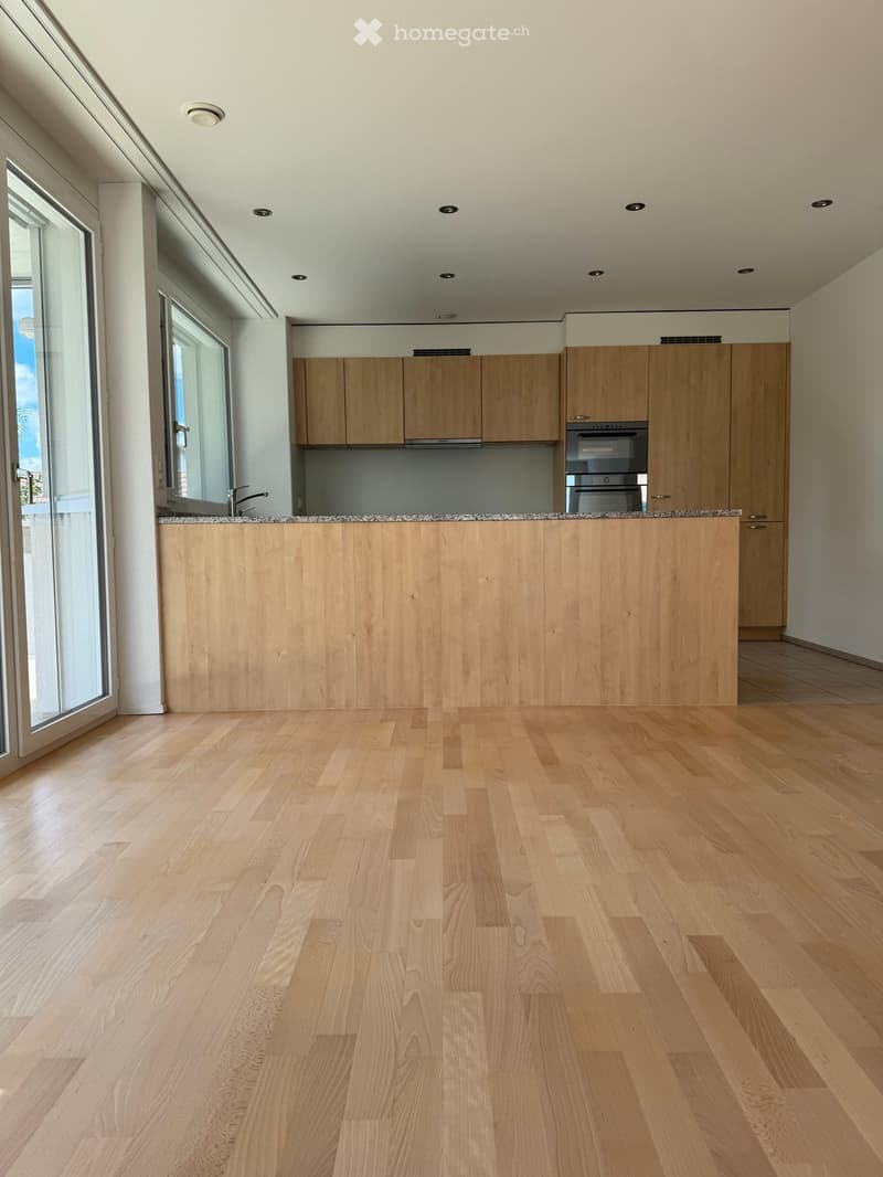 Empty kitchen with light wooden cabinets, a granite countertop, and a wooden floor. Large windows and a sliding door provide natural light.
