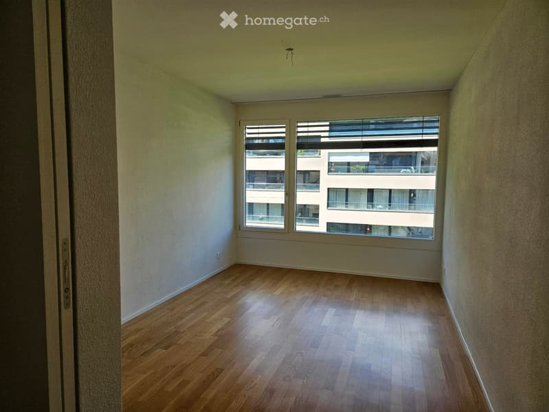 Empty room with wooden flooring, white walls, and large windows showing a view of neighboring buildings.