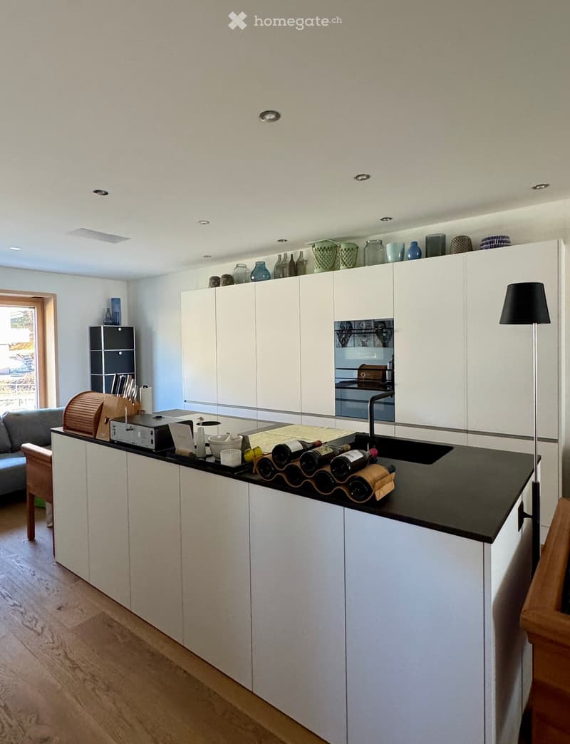 modern kitchen with large black island, white cabinets, wooden floors, black countertops, built-in oven, sink with faucet, wine rack on island, lamps, shelves with decorative items, gray sofa and coffee table